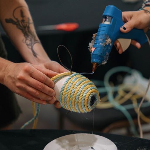 A hand holds a glue gun while another set of hands holds a buoy and wraps it in yellow and blue rope. 